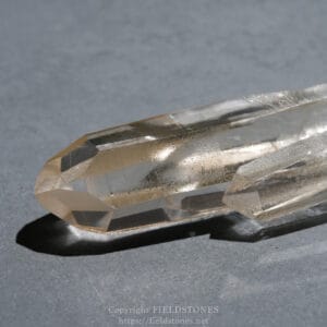Close-up of a clear, faceted quartz crystal point resting on a gray surface, with sharp beveled facets and a soft shadow