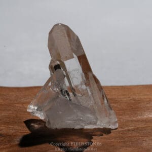 Clear quartz crystal cluster standing on a wooden surface, with sharp faceted prisms catching light/shine on the wood table.