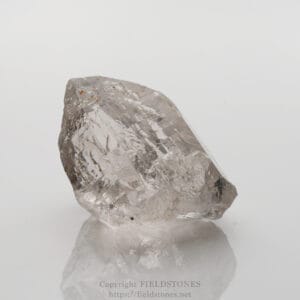 Clear quartz crystal shard on a white reflective surface, showcasing facets and natural striations with subtle gray hues.