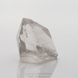 Clear quartz crystal prism on a white reflective surface, showing faceted faces and natural inclusions.