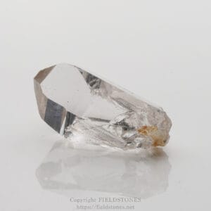 Clear quartz crystal with faceted, transparent body resting on a reflective surface, showing a rough, yellowish base edge.