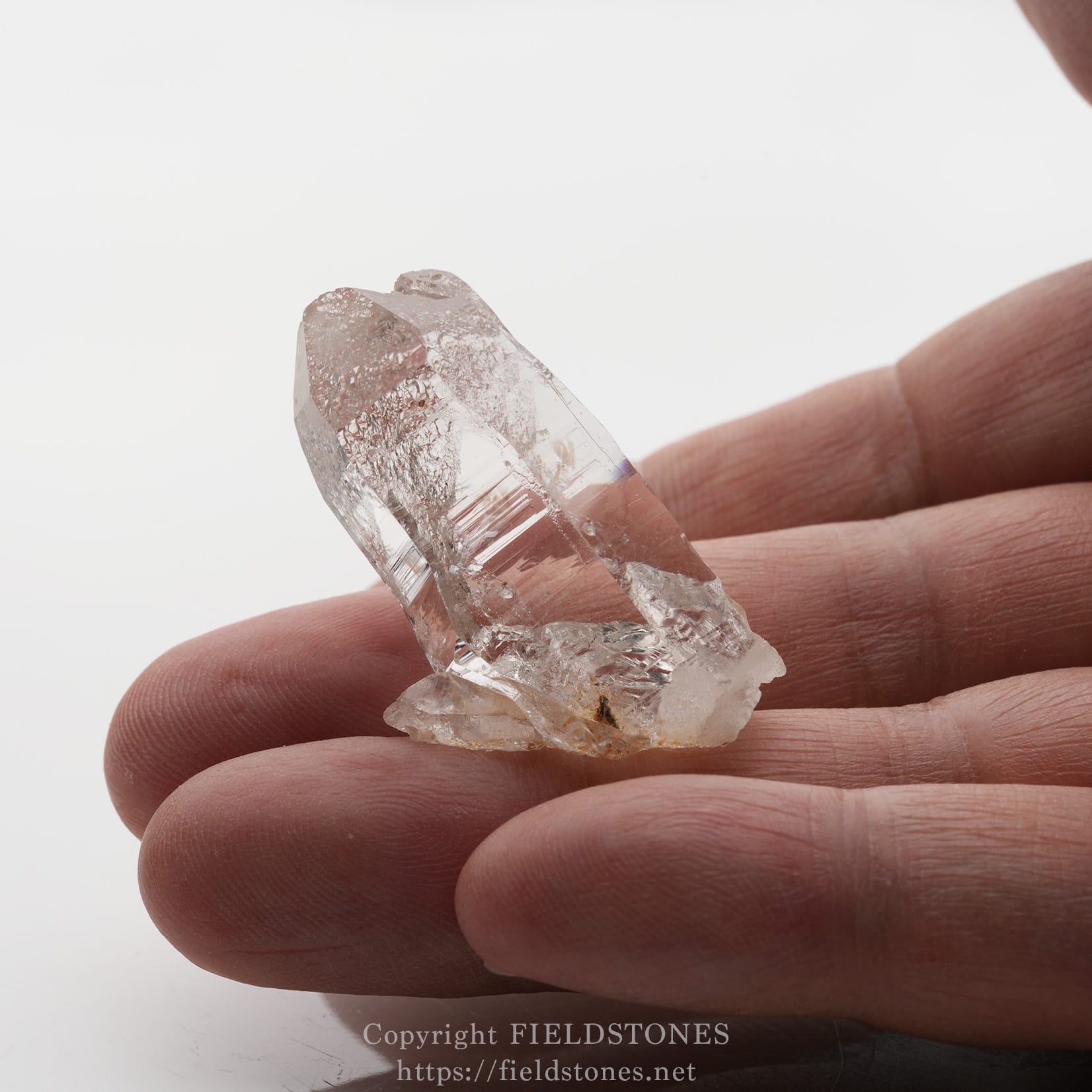 6501-12 - FieldStones Clear quartz crystal held between fingertips, showing faceted, transparent surfaces against a white background.