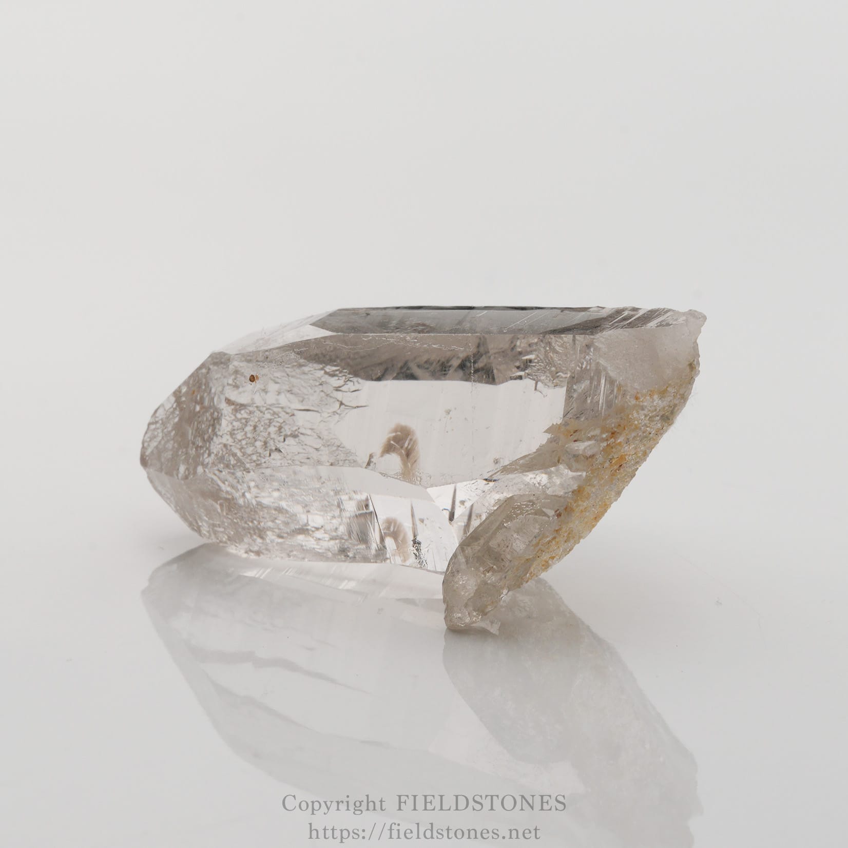 6501-3 - FieldStones Clear quartz crystal shard resting on a white, reflective surface, showing transparent facets and a rough base.