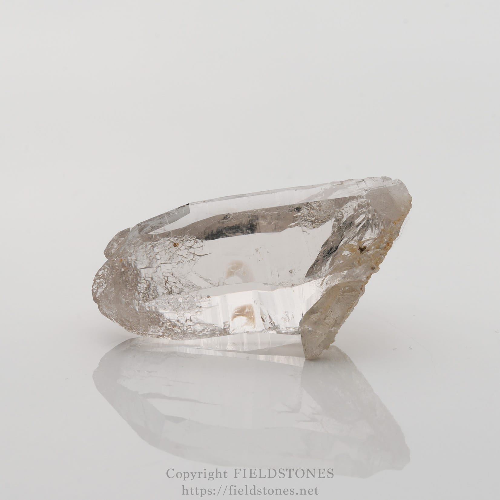 6501-5 - FieldStones Clear quartz crystal with rough edges resting on a white reflective surface.
