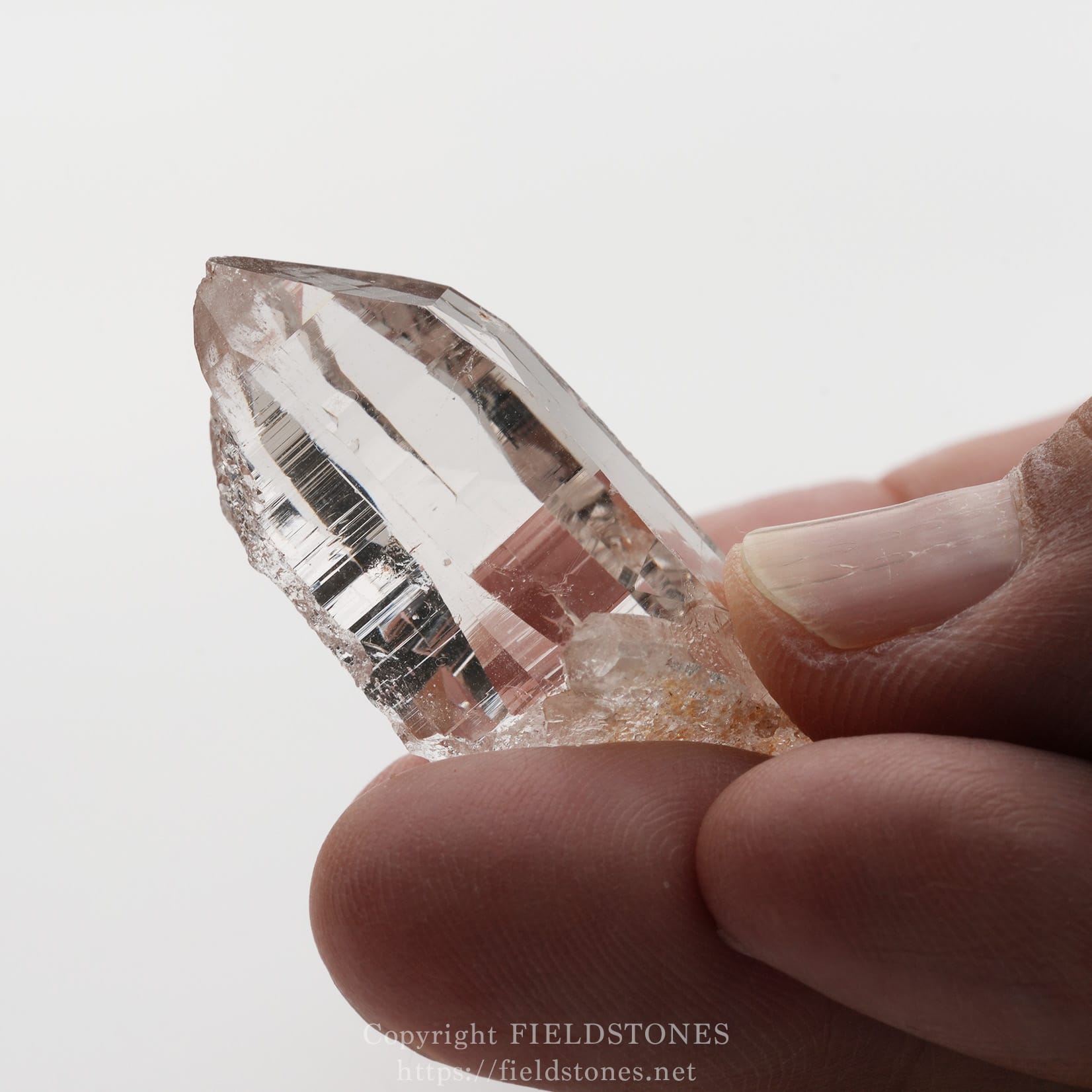 6501-7 - FieldStones Close-up of a clear quartz crystal held between a person's thumb and index finger against a pale background instructive image of a mineral sample.