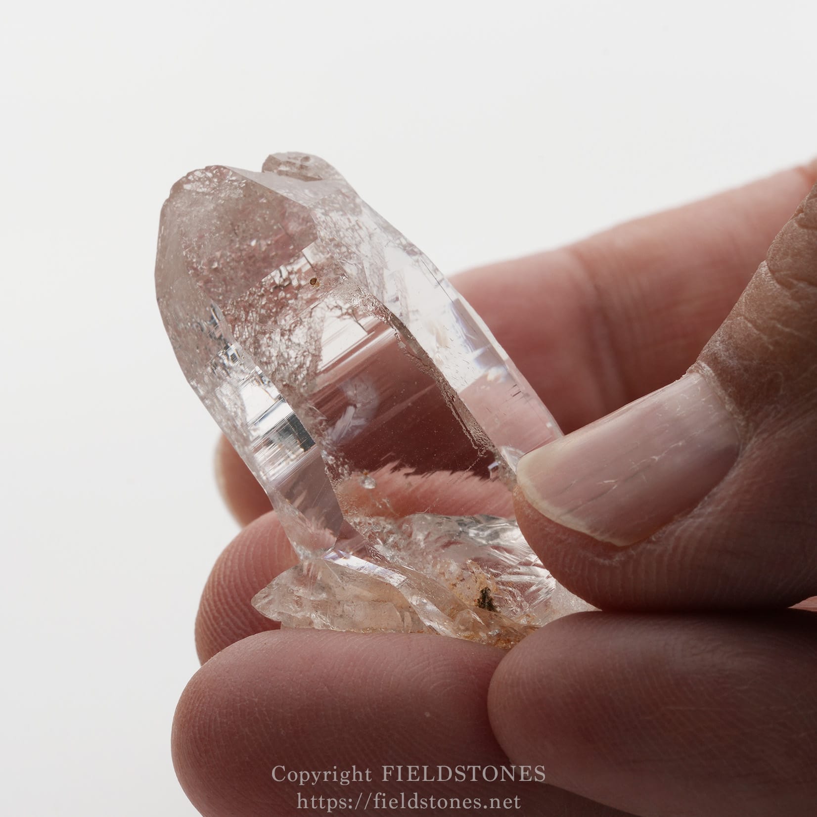 6501-9 - FieldStones Close-up of a clear quartz crystal point being held between fingertips, showing natural facets and a few internal inclusions.