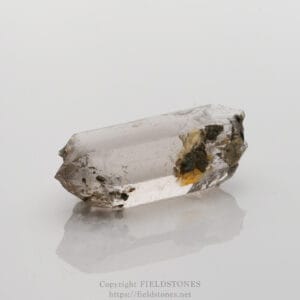 Clear quartz crystal with dirt and yellow inclusions on a white reflective surface.