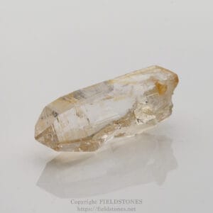 Clear quartz crystal specimen lying on a white surface with subtle reflection and pale yellow inclusions nearby
