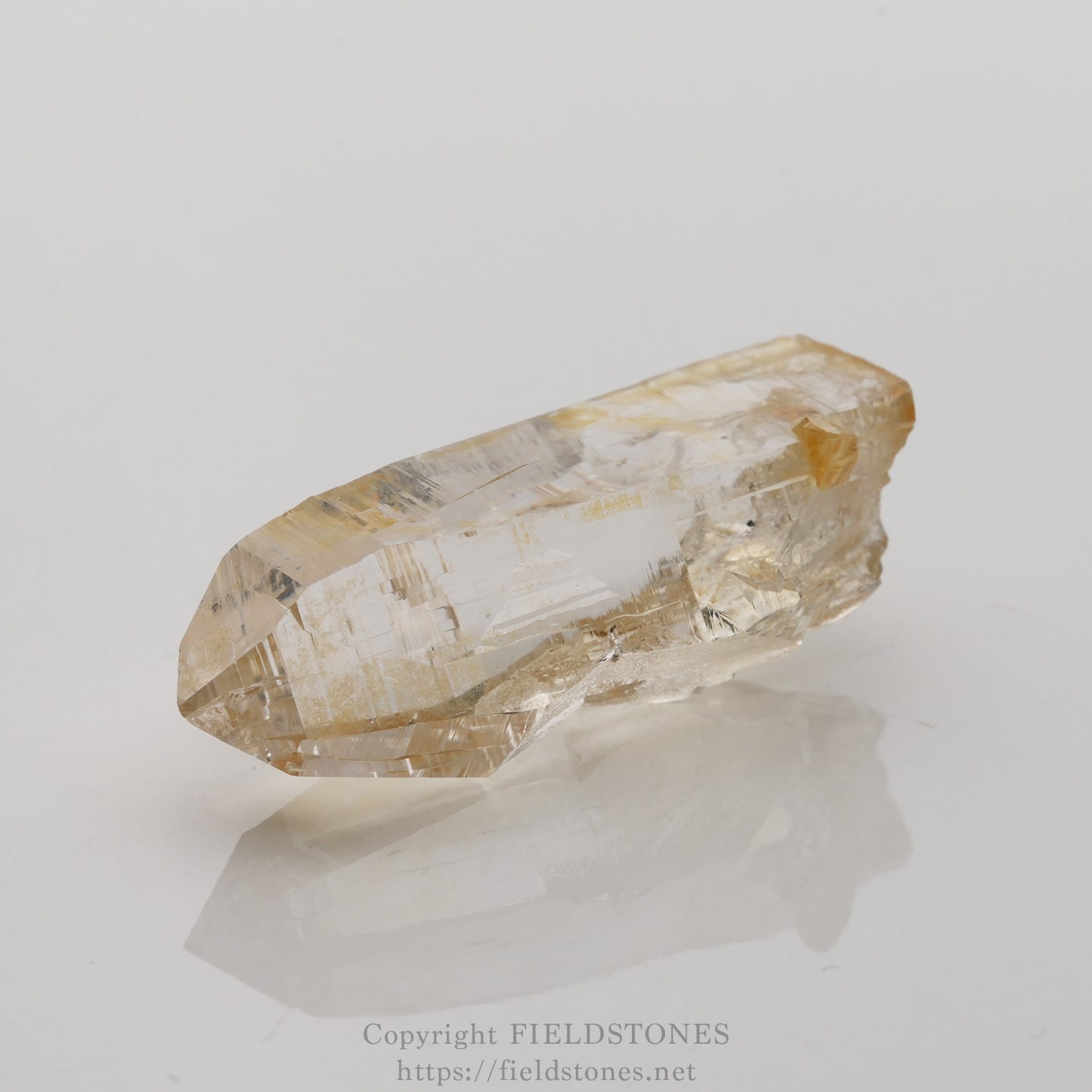 6506-1 - FieldStones Clear quartz crystal specimen lying on a white surface with subtle reflection and pale yellow inclusions nearby