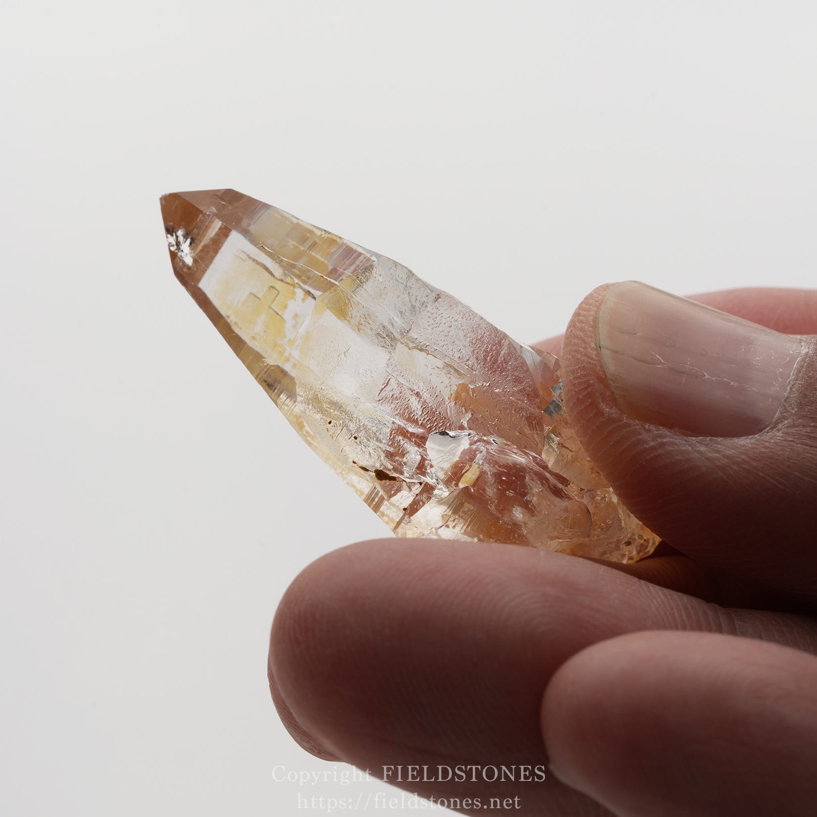 6506-10 - FieldStones Clear quartz crystal with a pointed tip held between two fingers against a white background.