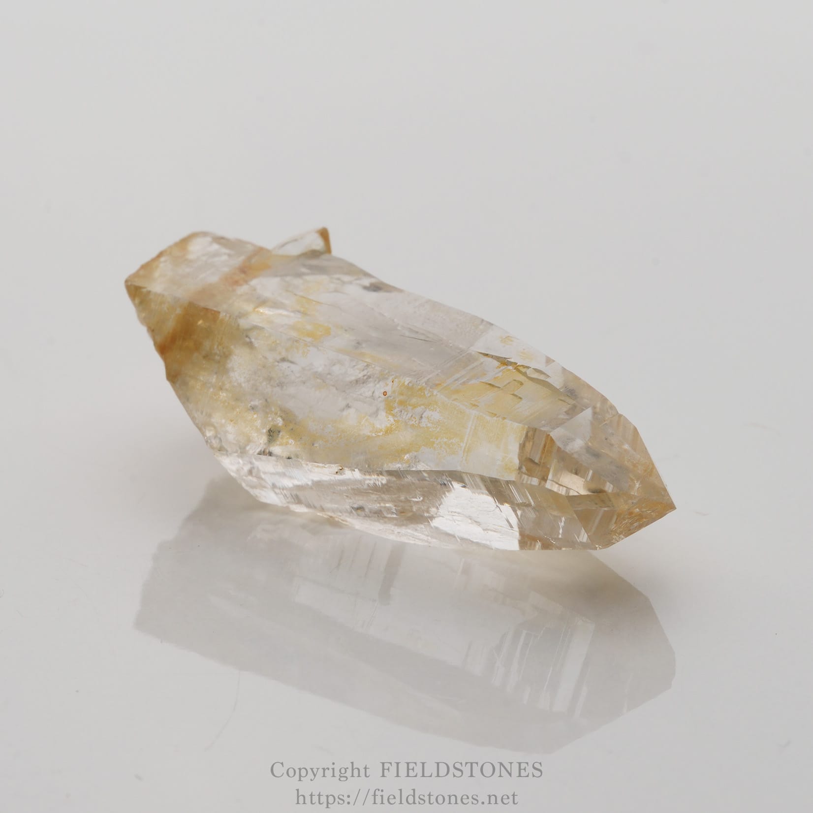 6506-2 - FieldStones Clear quartz crystal point with pale yellow inclusions on a white reflective surface.