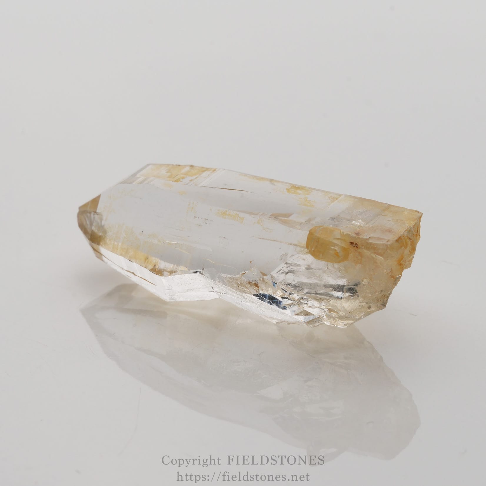 6506-3 - FieldStones Clear quartz crystal with pale yellow inclusions, photographed on a clean white surface; a natural, elongated hexagonal prism with a pointed termination on one end.
