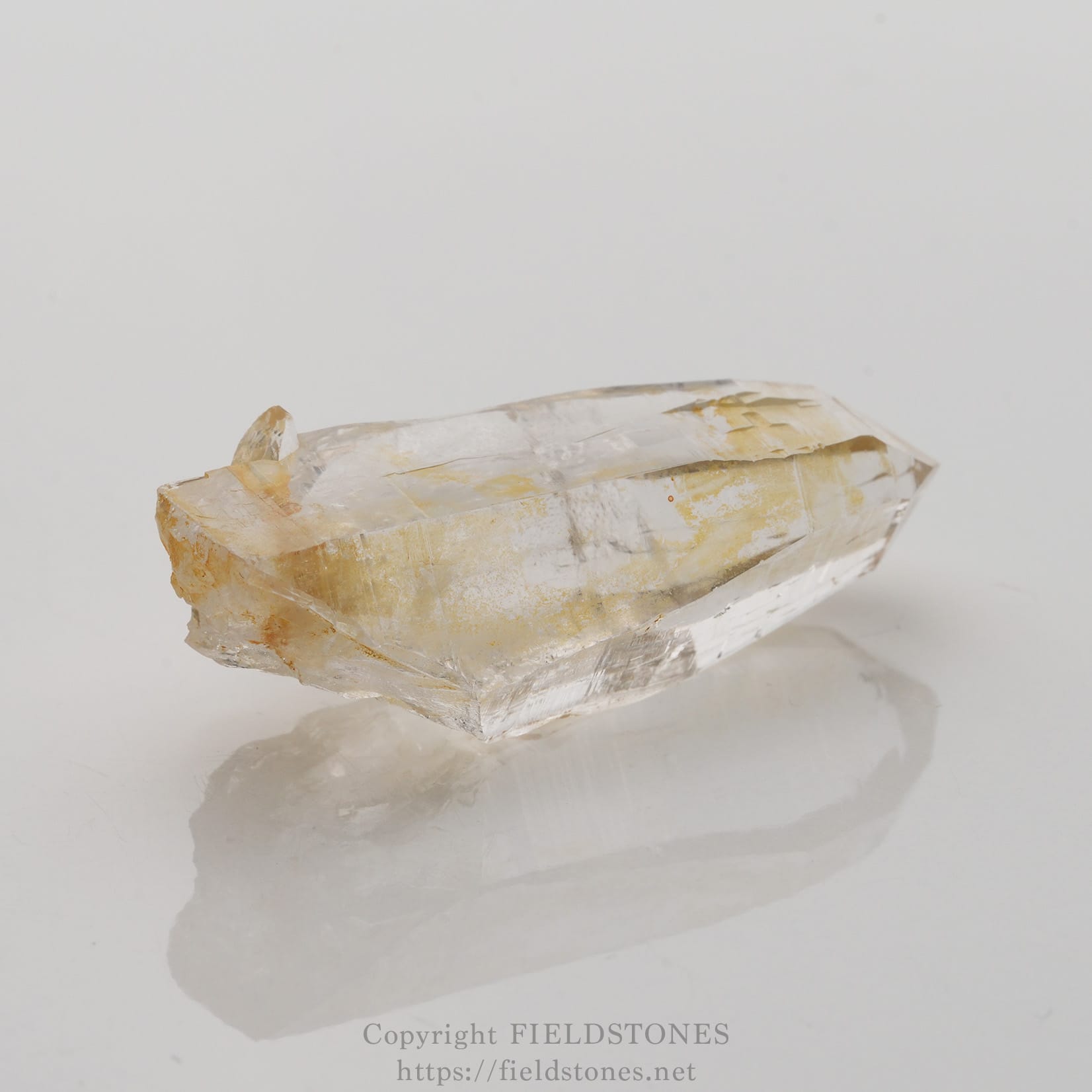 6506-4 - FieldStones Clear quartz crystal point with pale yellow inclusions on a glossy white surface.