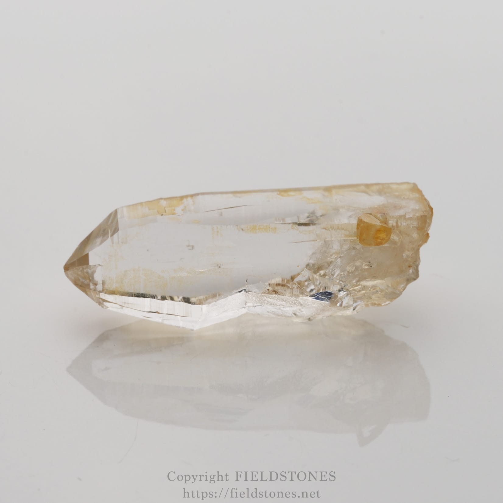 6506-5 - FieldStones Clear quartz crystal with a small yellow inclusion, lying on a white reflective surface.
