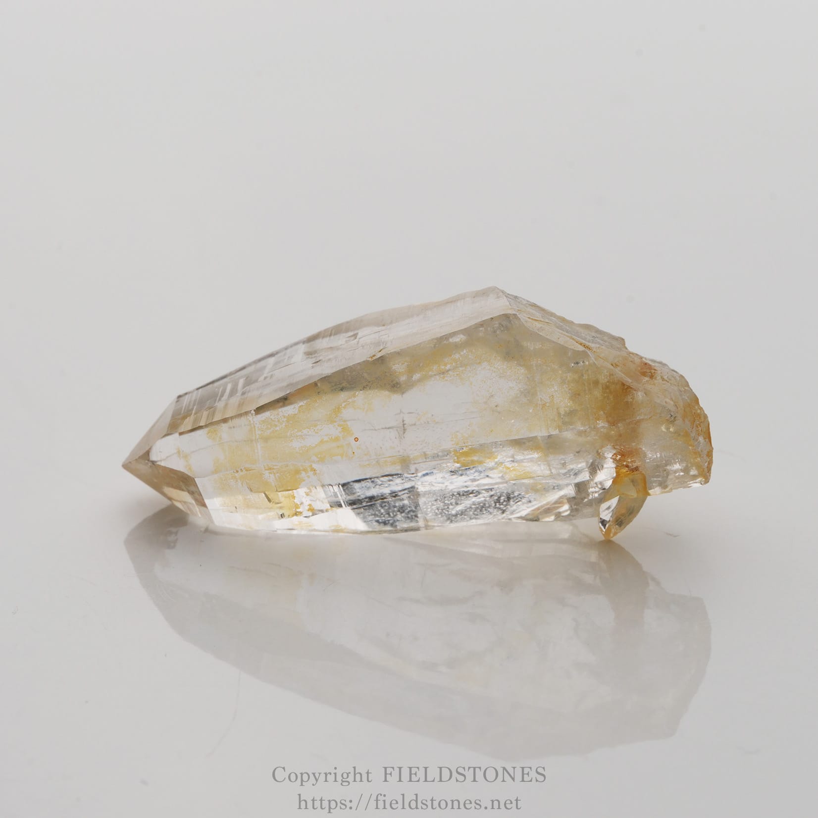 6506-7 - FieldStones Clear quartz crystal specimen with pale yellow inclusions resting on a white surface, reflecting below.
