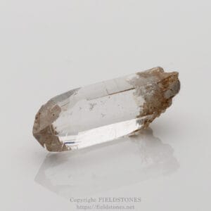 Transparent quartz crystal point with a rough brown matrix at one end, resting on a white, reflective surface.