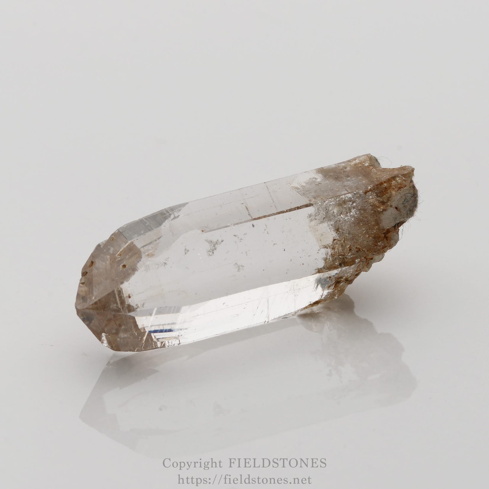 6508-1 - FieldStones Transparent quartz crystal point with a rough brown matrix at one end, resting on a white, reflective surface.