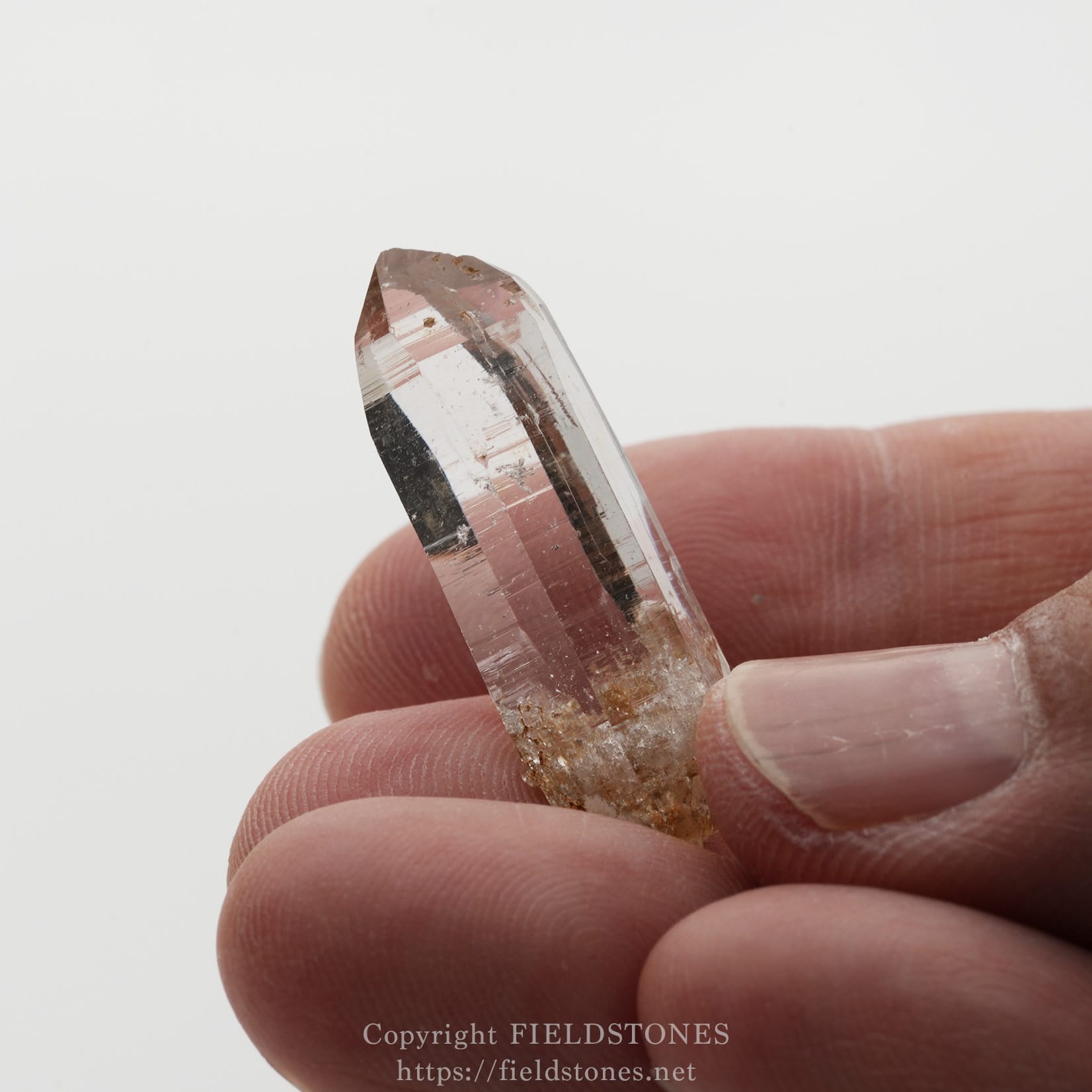 6508-12 - FieldStones Clear quartz crystal point being held between a thumb and forefinger, showing a faceted termination and transparent body.