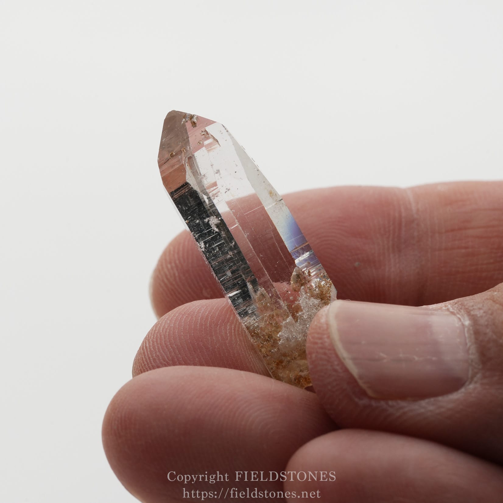 6508-13 - FieldStones Close-up of a transparent quartz crystal point held between fingertips, showing clear facets and a pinkish tint with some brownish mineral inclusions.