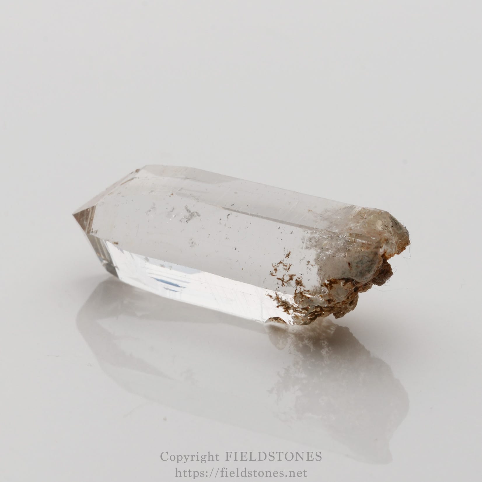 6508-2 - FieldStones Clear quartz crystal prism with a rough, brownish base resting on a reflective white surface.