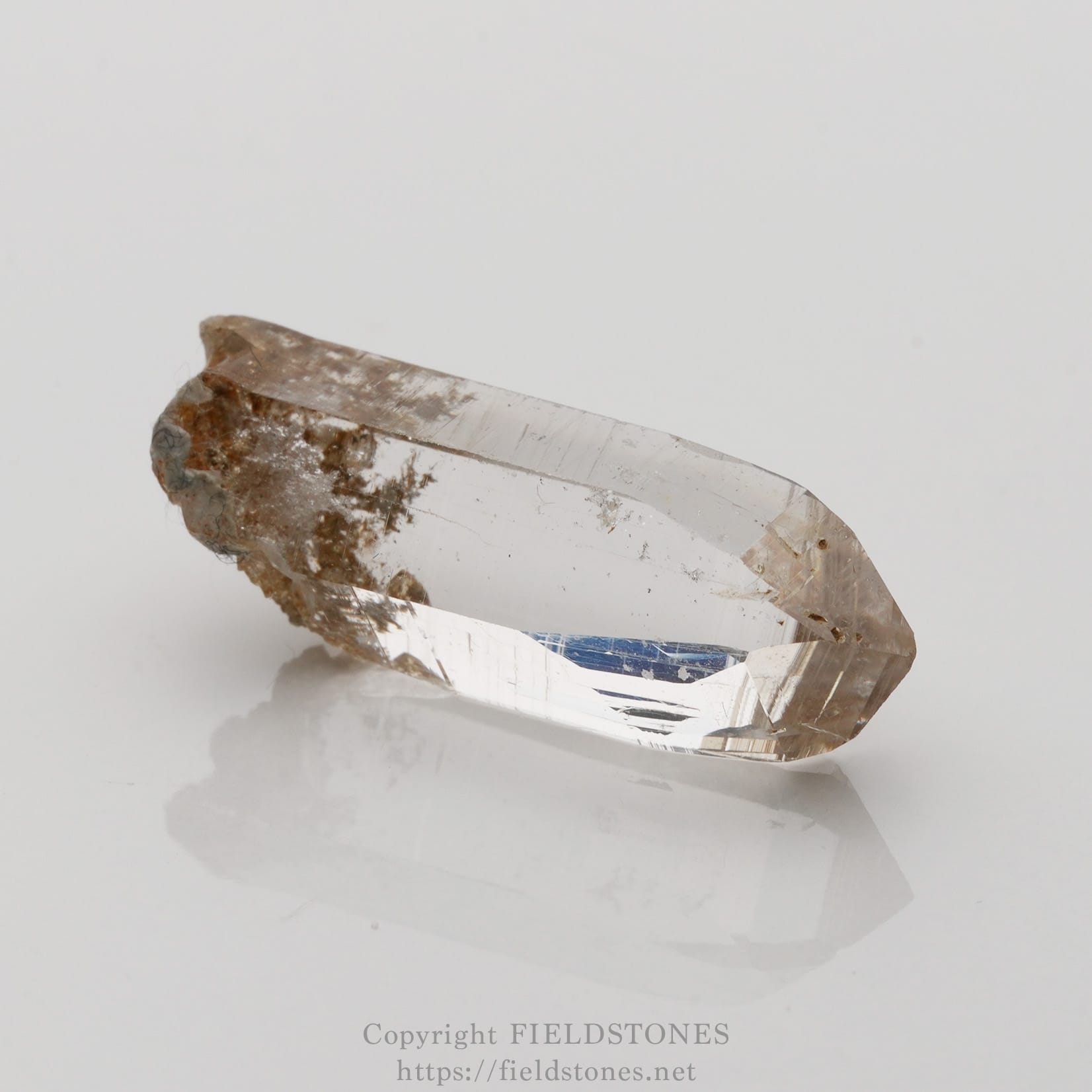 6508-4 - FieldStones Clear quartz crystal point with brown matrix on a white surface, photographed as a mineral specimen.
