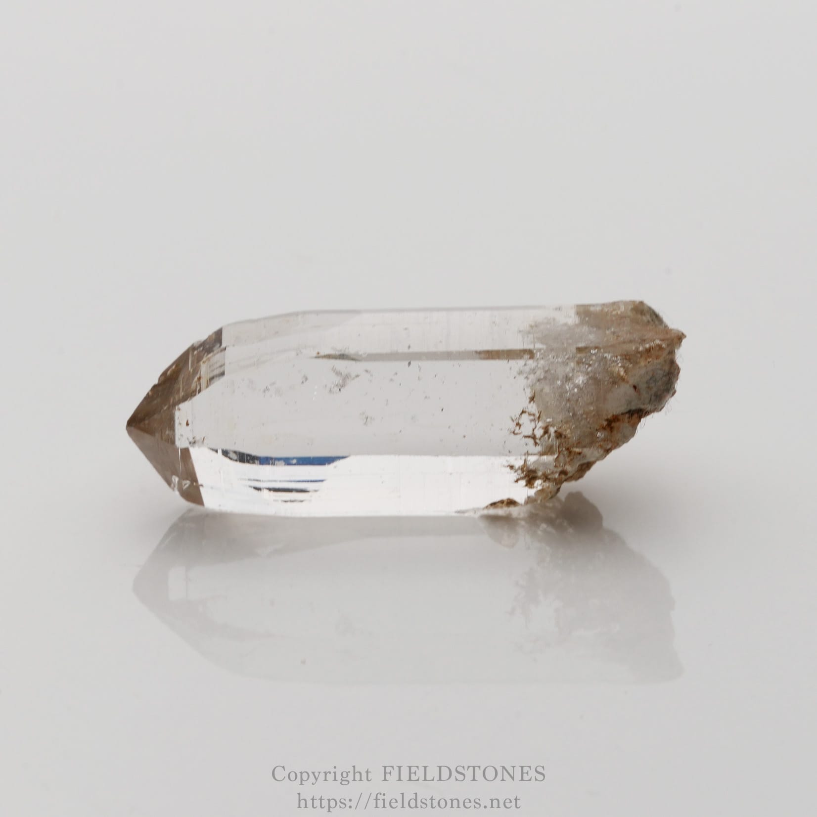 6508-5 - FieldStones Clear quartz crystal with brown inclusions, resting on a white reflective surface.