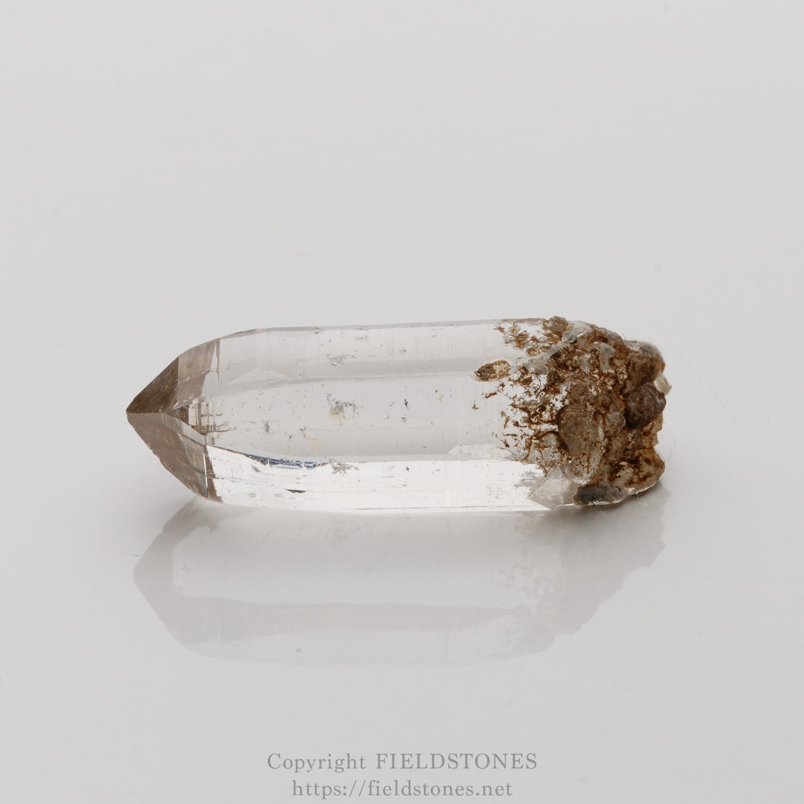6508-6 - FieldStones Clear quartz crystal point with a brown rock matrix on one end, lying on a white surface.