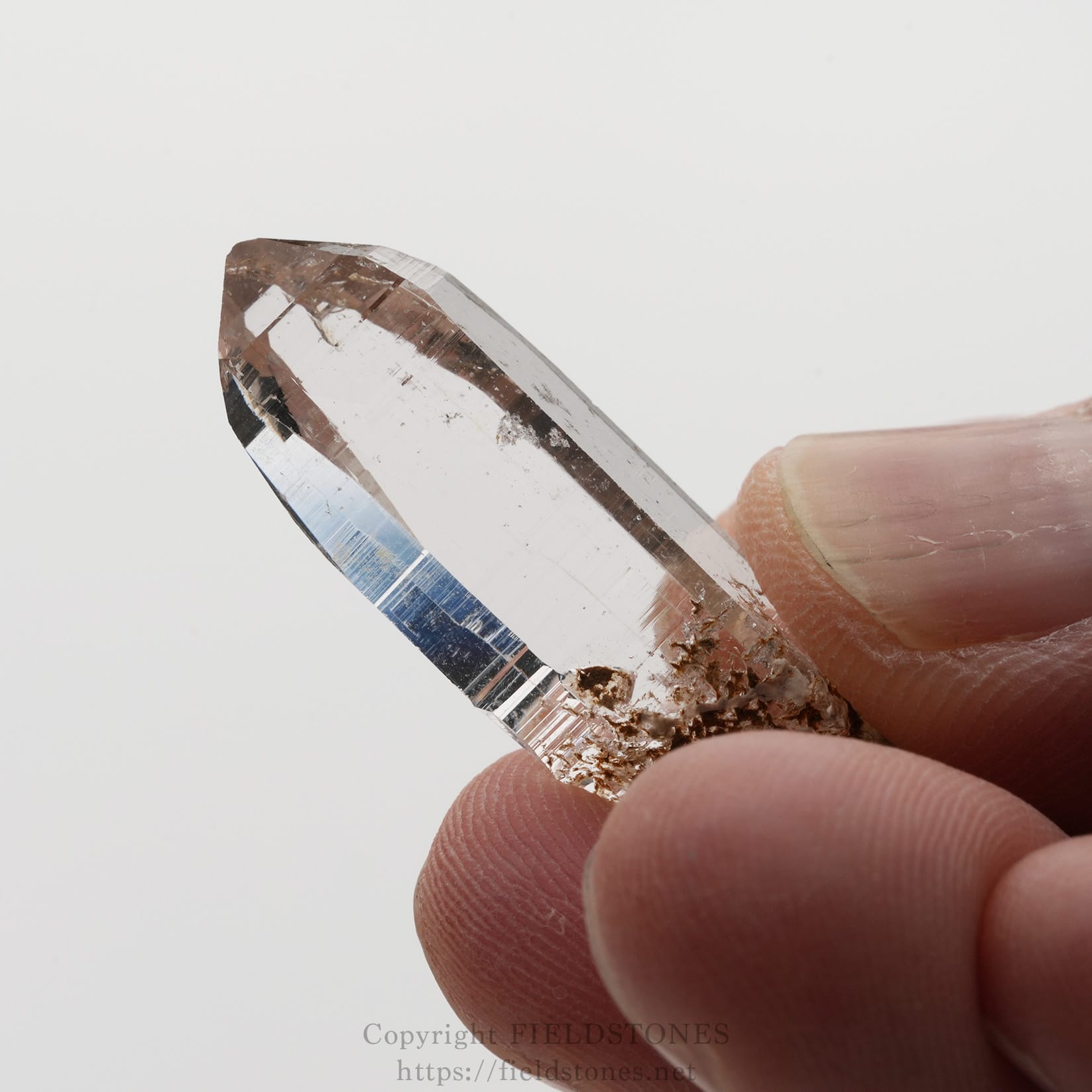 6508-8 - FieldStones Clear quartz crystal point held between fingertips, showing a sharp, elongated prism with a brown mineral base at the bottom.