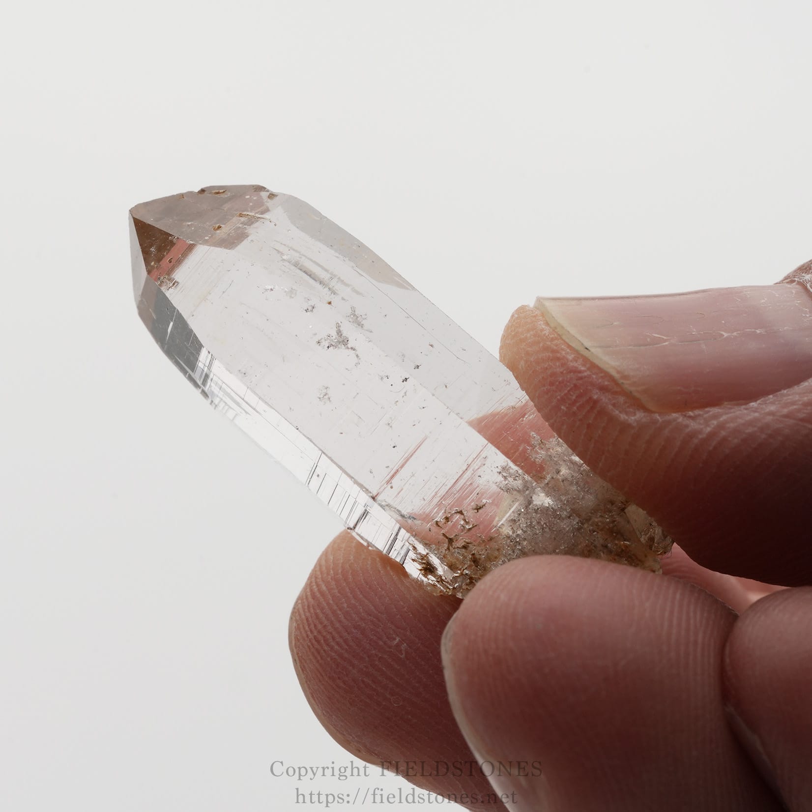 6508-9 - FieldStones Clear quartz crystal point held between a thumb and forefinger against a plain background.