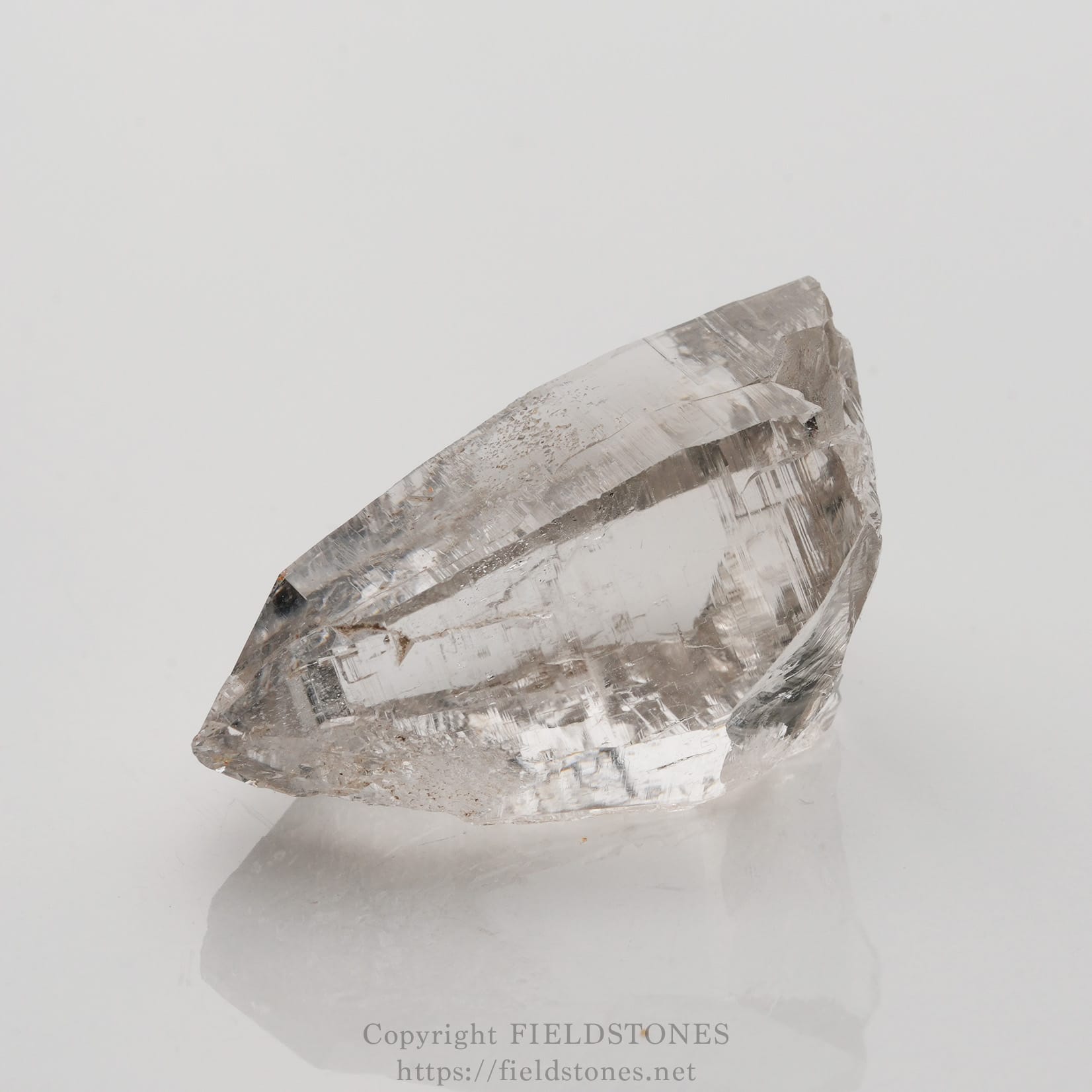 6509-1 - FieldStones Clear quartz crystal point with natural facets resting on a white reflective surface.
