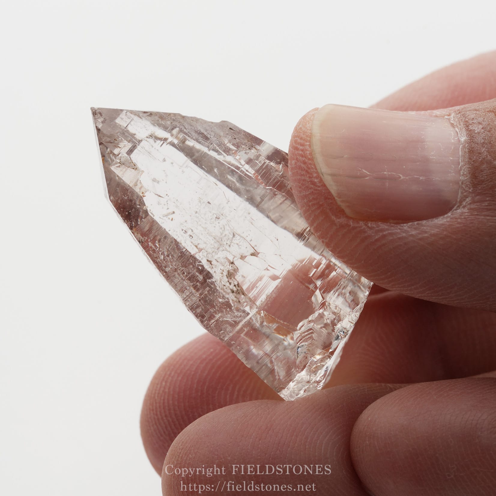 6509-10 - FieldStones Close-up of a clear crystal shard held between a thumb and finger, showing sharp facets and translucence over a white background.