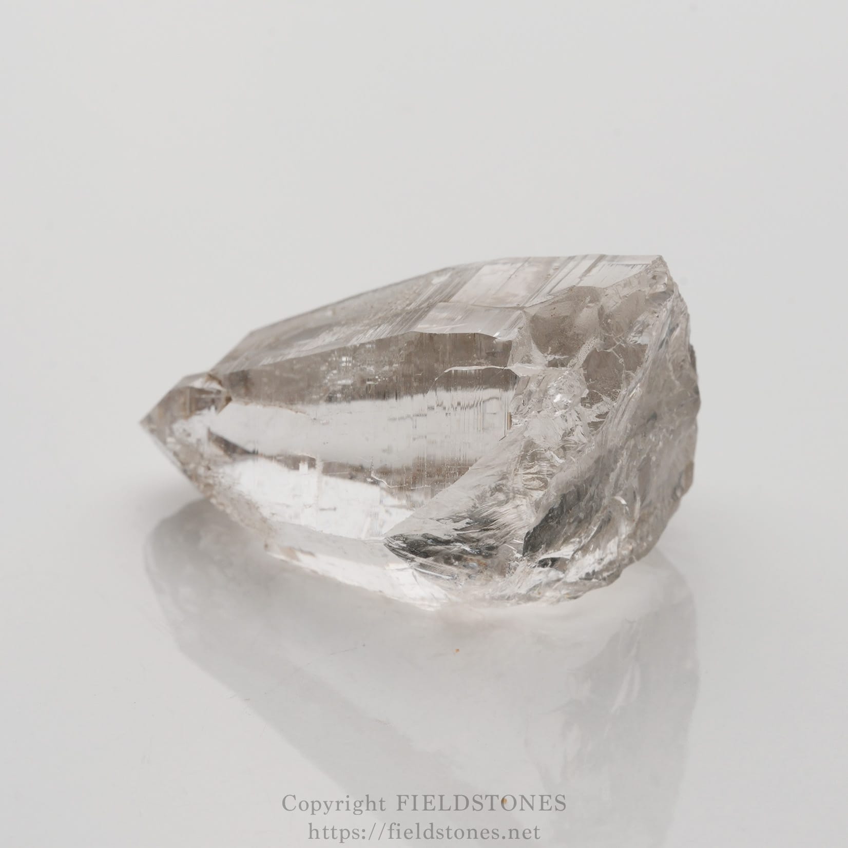 6509-2 - FieldStones Clear quartz crystal with a sharp pointed tip resting on a reflective white surface.