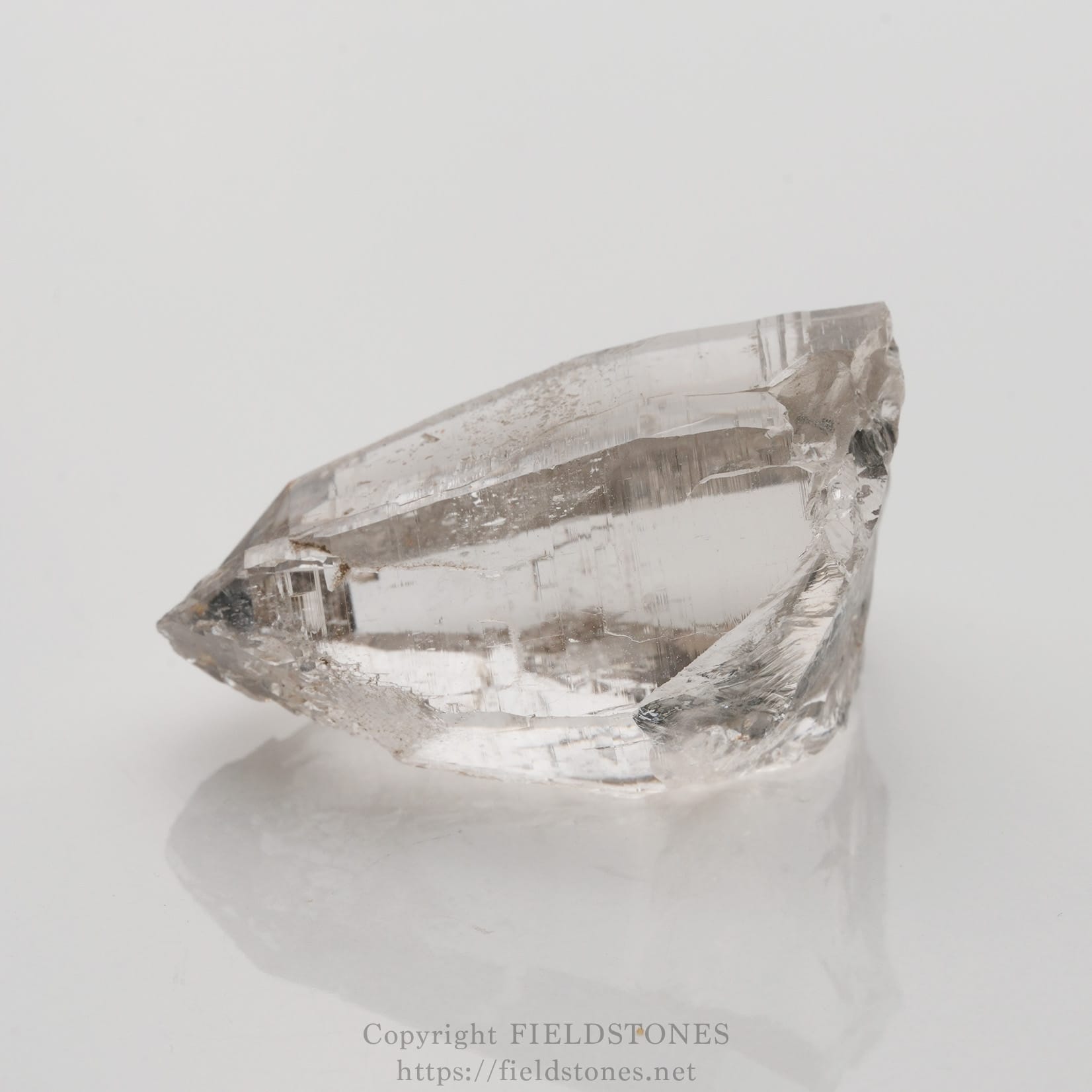 6509-5 - FieldStones Clear quartz crystal point with sharp tip resting on a smooth white surface, reflective underneath