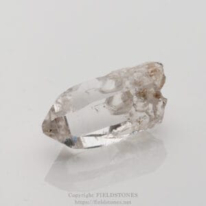 Clear quartz crystal with a natural, rough base resting on a white surface.