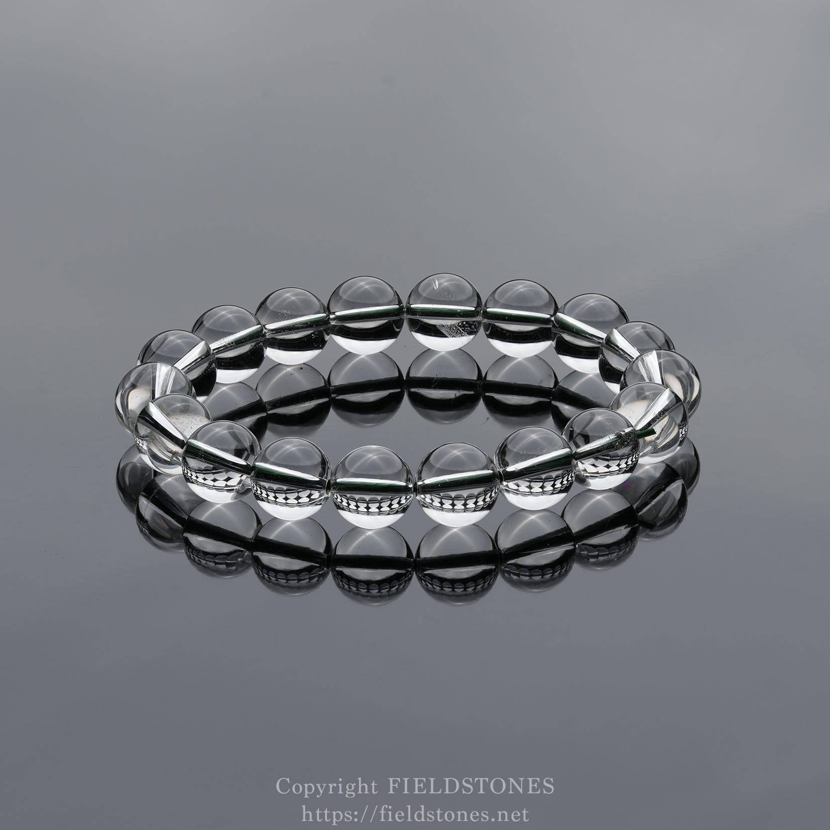 6512-1 - FieldStones Clear glass bead bracelet arranged in a circular loop on a glossy reflective surface, showcasing smooth, transparent beads.