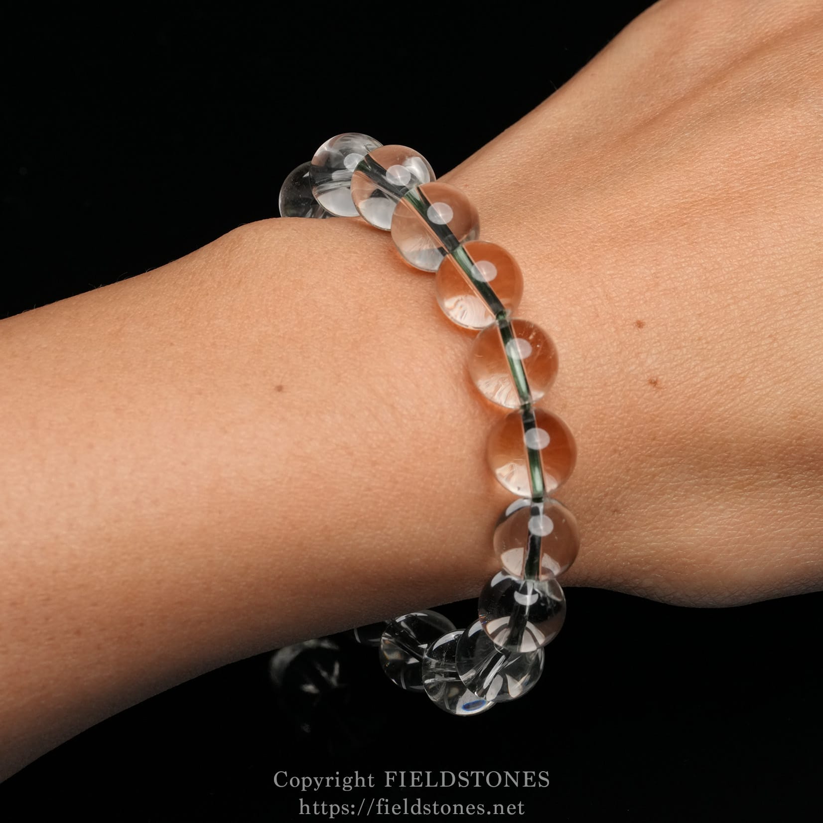 6512-13 - FieldStones Transparent glass bead bracelet on a wrist, with round clear beads and a green thread showing between beads on a black background.