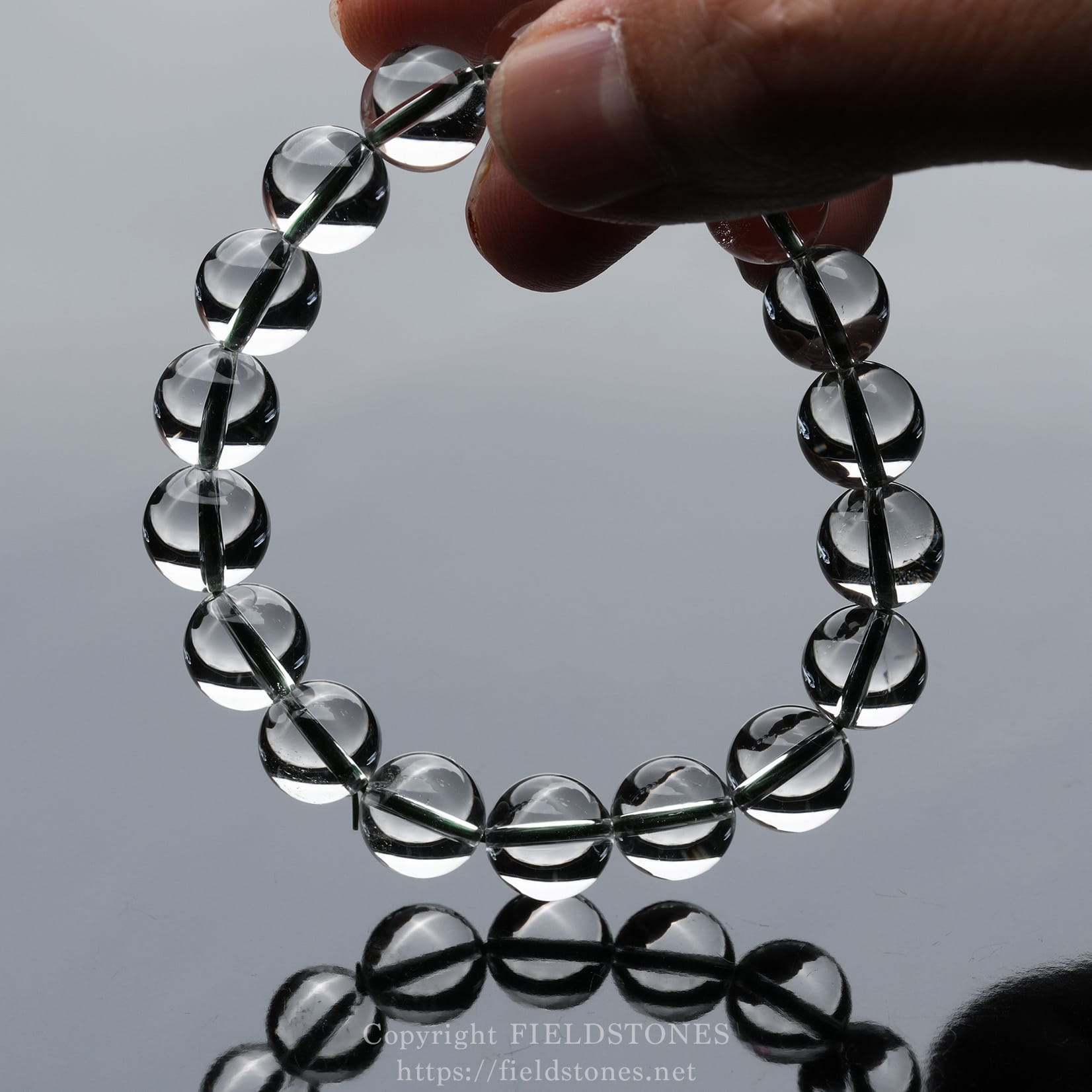6512-3 - FieldStones Clear glass bead bracelet formed into a circle, held by fingers over a mirrored surface.