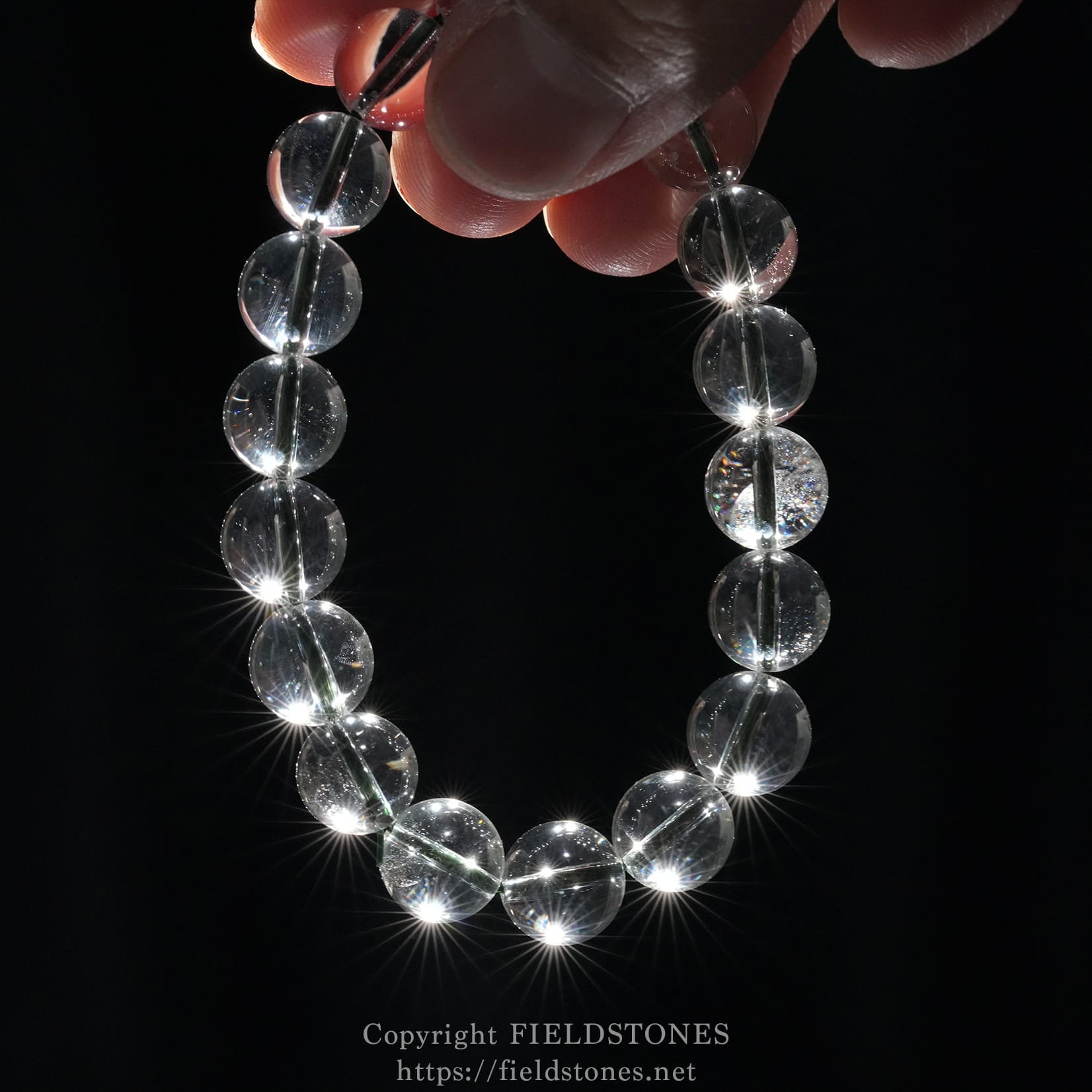 6512-6 - FieldStones Hand holding a strand of transparent glass beads sparkling against a black background.