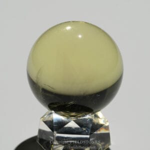 Two-tone sphere (stone) with a pale yellow upper half and dark lower half resting on a clear faceted crystal stand.