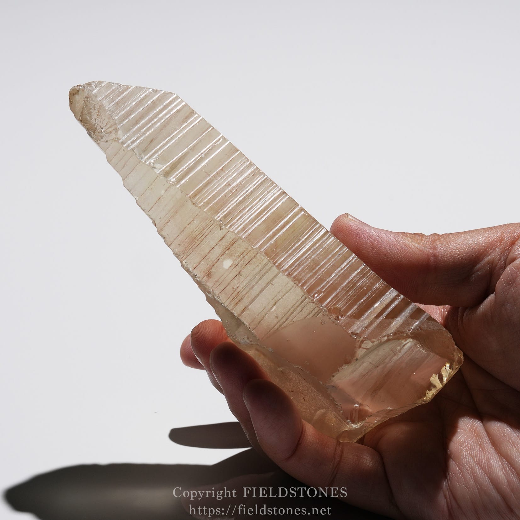 6524-10 - FieldStones Pinkish translucent quartz crystal blade held between thumb and fingers, showing parallel striations along its length.