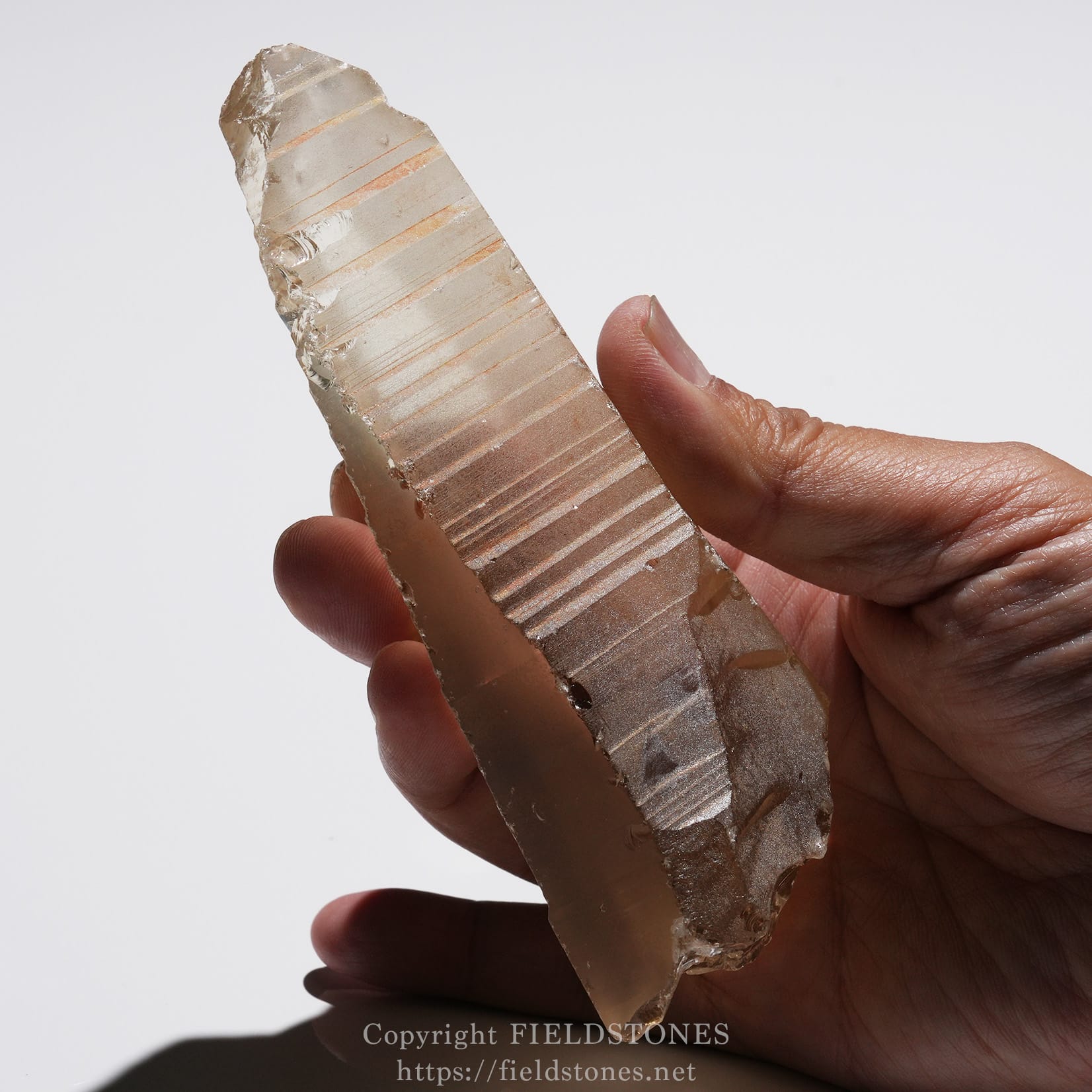 6524-14 - FieldStones Hand holding a translucent quartz crystal wand with horizontal striations against a light background.