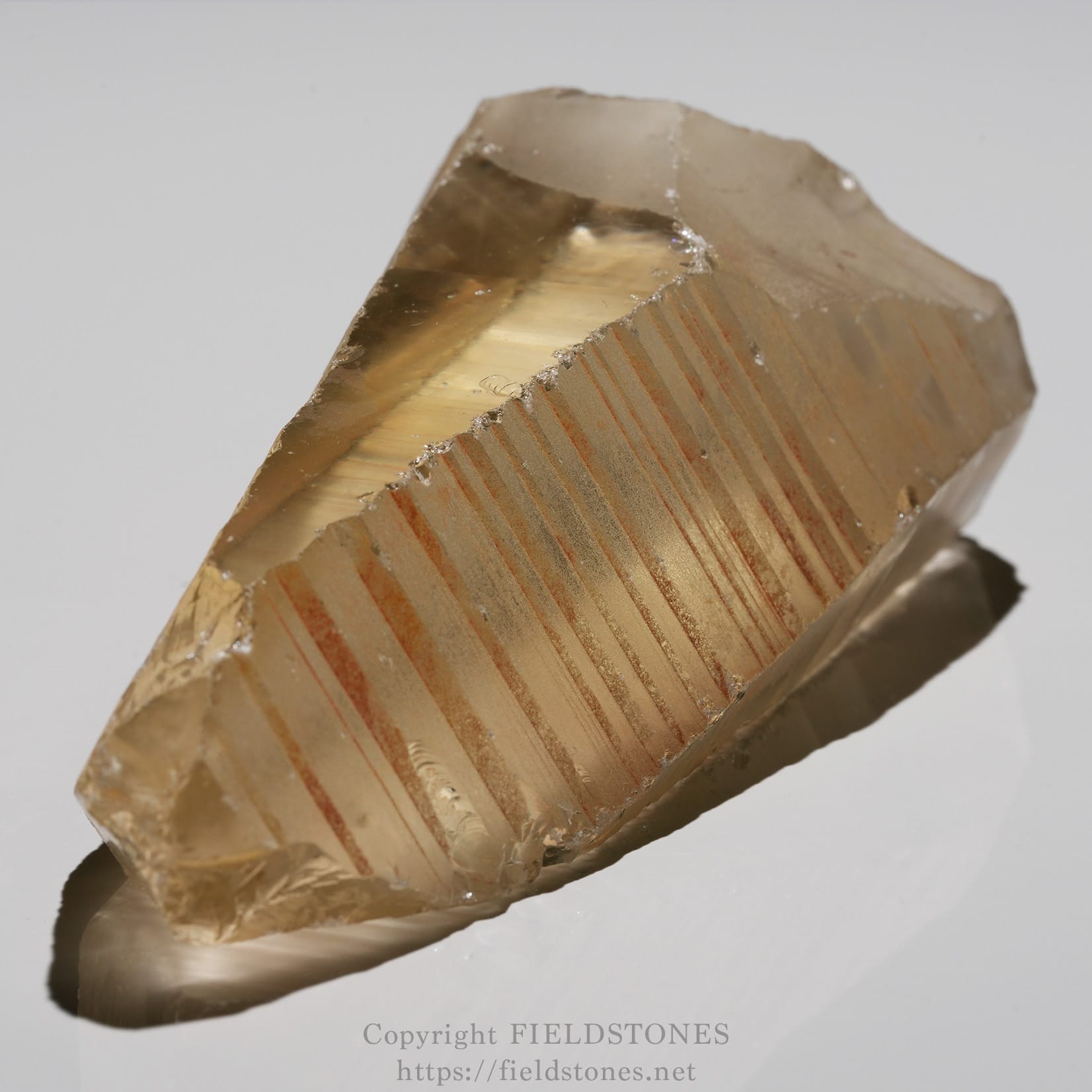 6524-3 - FieldStones Golden translucent mineral shard with parallel reddish-brown striations on one face, resting on a reflective surface.
