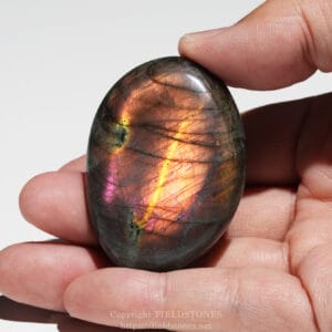 Polished labradorite stone held between fingertips, displaying iridescent rainbow colors.
