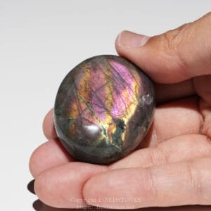 Polished labradorite stone held between fingers showing iridescent pink, purple, and gold flashes.