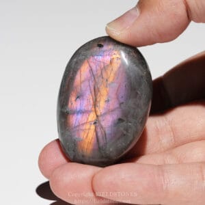 Close-up of a hand holding a polished labradorite stone with iridescent blue, purple, and orange flashes across its surface.