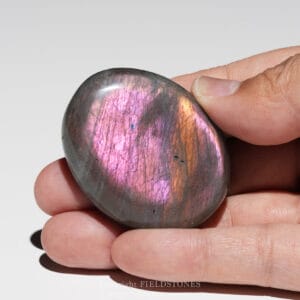 Hand holding a polished, iridescent purple-pink stone (likely labradorite or similar) against a plain background.