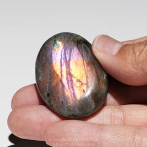 Polished labradorite stone showing iridescent rainbow colors (blue, pink, gold) held between fingertips by a person’s hand.