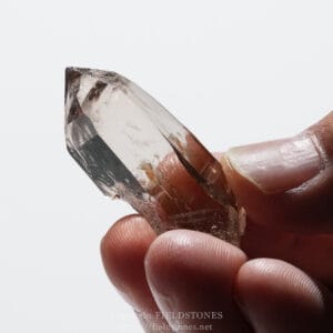 Close-up of a faceted, transparent crystal held between a thumb and index finger against a white background.
