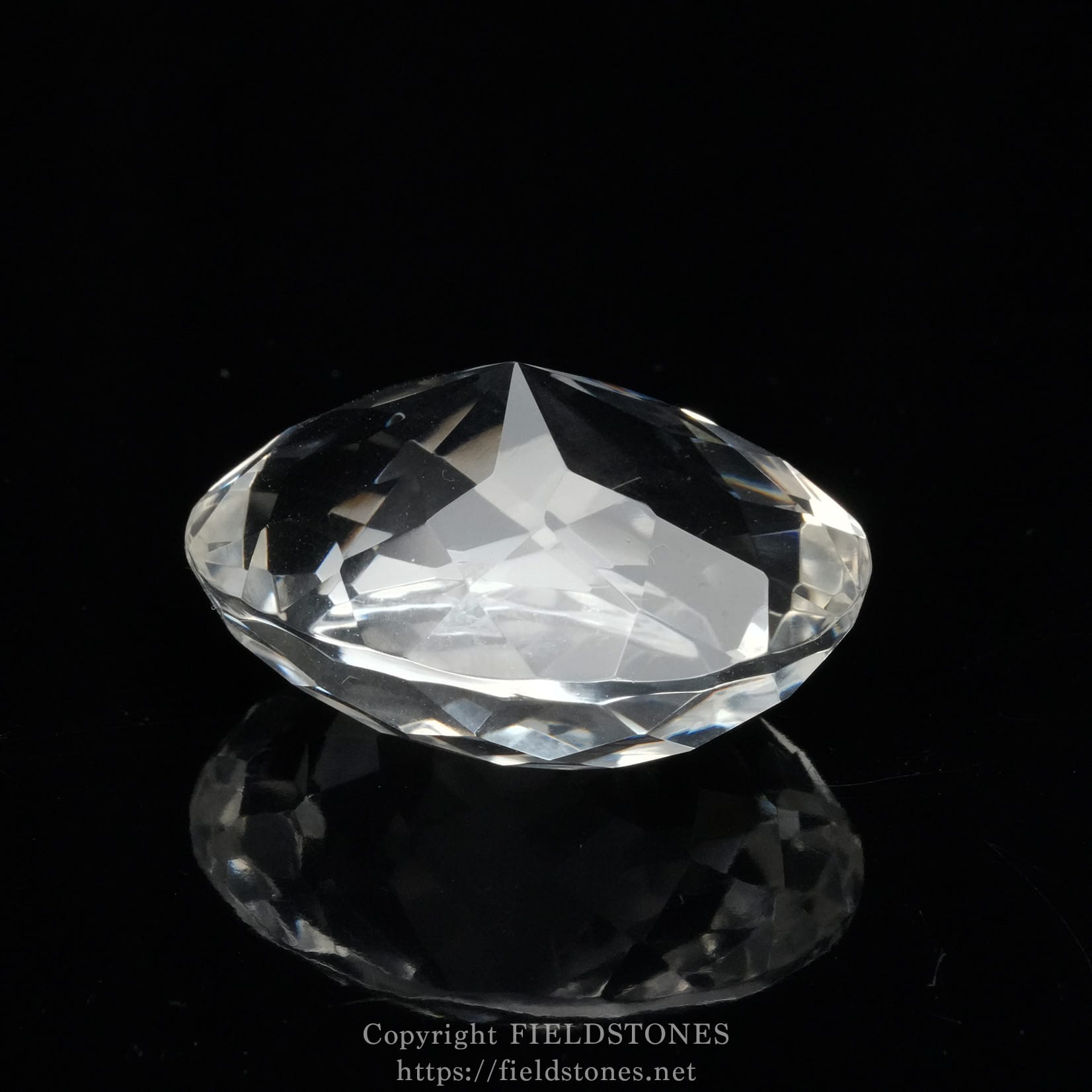 6537-14 - FieldStones Diamond-shaped crystal gemstone resting on a glossy black surface, facets sparkling under light with a faint reflection beneath.