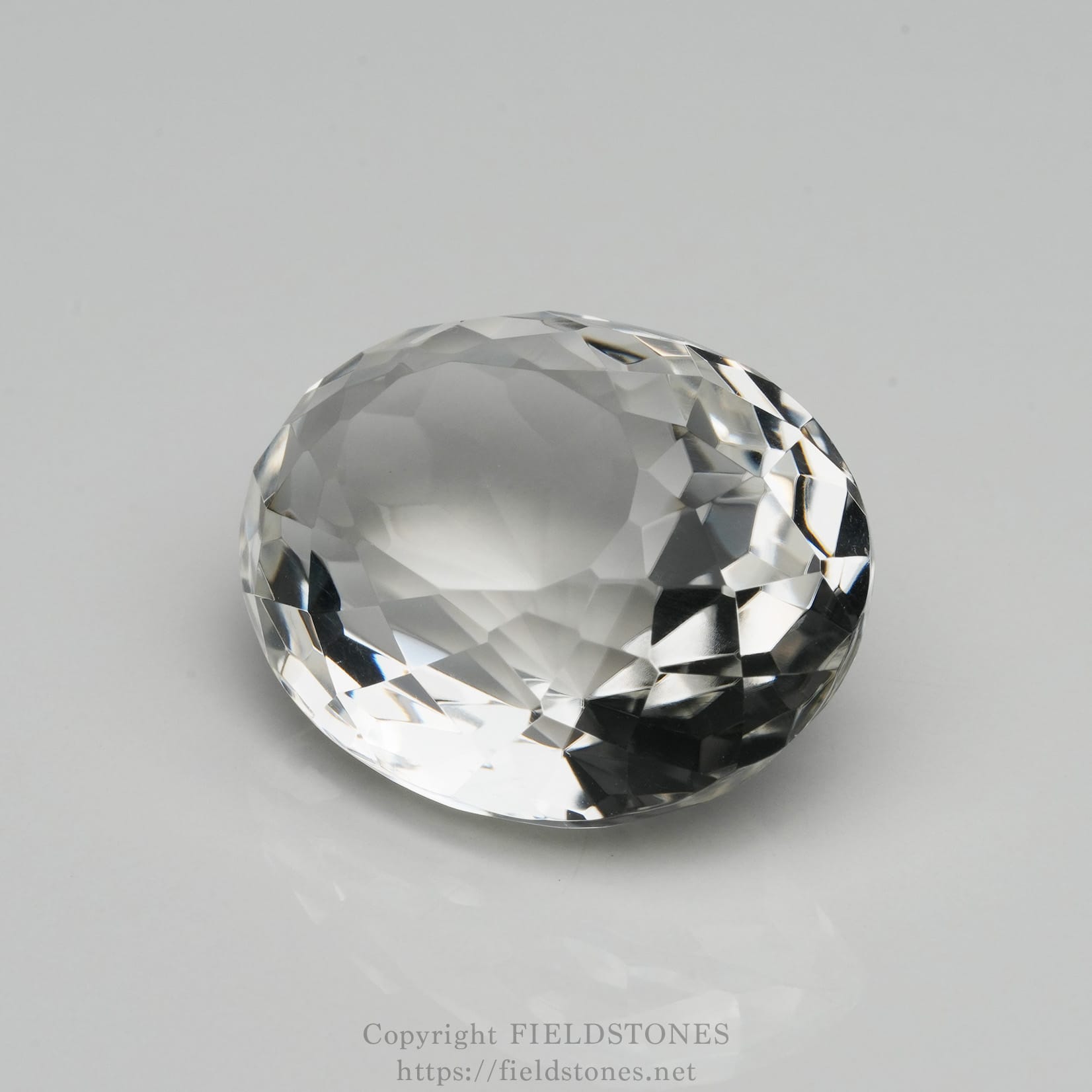 6537-2 - FieldStones Close-up of a faceted clear crystal gemstone resting on a light gray surface, showing sparkling facets and reflections.